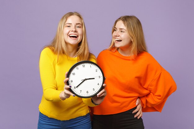 Two cheerful young blonde twins sisters girls in vivid colorful clothes hold round clock isolated on pastel violet blue wall. People family lifestyle concept. 