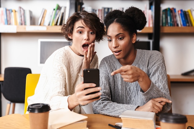 Two cheerful young girls students studying at library, taking a selfie