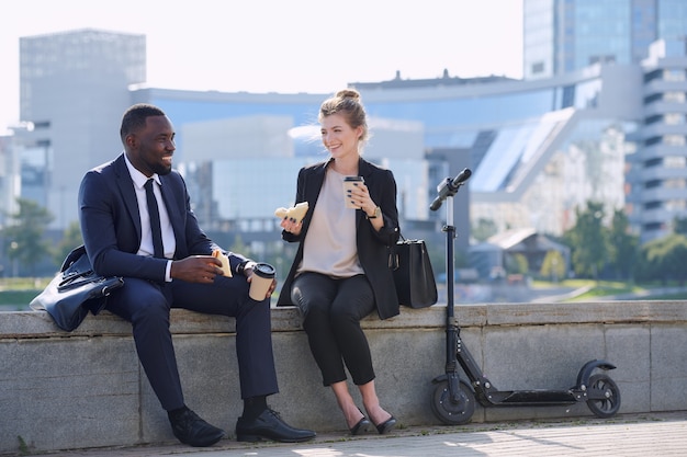 Two cheerful young intercultural business partners having coffee and tasty sandwiches while enjoying break by riverside in urban environment