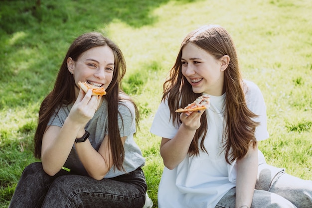 Two cheerful young teen friends in the park eating pizza. Women eat fast food. Not a healthy diet. Soft selective focus.