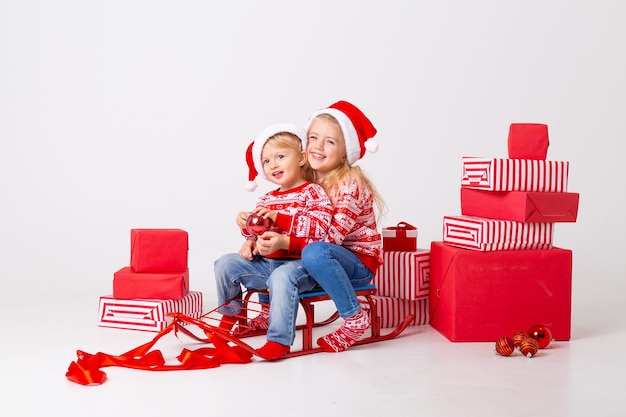 Two children a boy and a girl in sweaters and hats santa sit on a sled to carry gifts. studio, white background, space for text