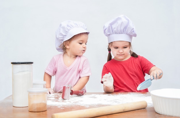 Two children in chef's hats are playing with flour. Elder girl shows the baby the action