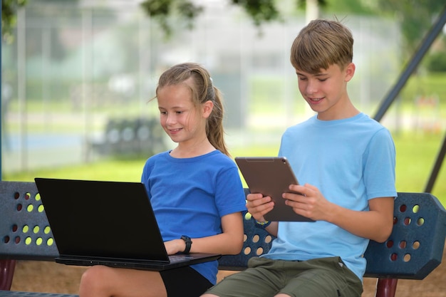 Two children teenager boy with laptop computer and young girl with digital tablet looking at screens outdoors sitting on bench in summer park Remote education during quarantine concept