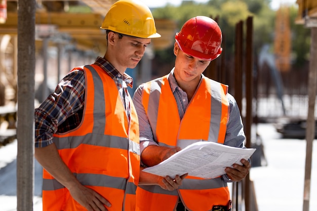 Two civil engineers dressed in orange work vests and helmets explore construction documentation on the building site near the wooden building constructions .