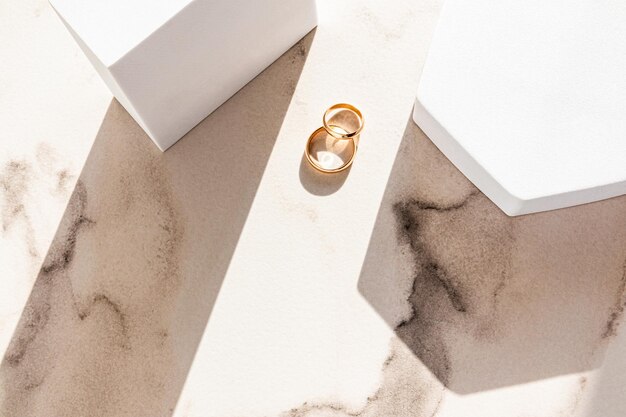 Two classic gold wedding rings lie on a marble beige background between two white geometric podiums of figures and sun rays Top view