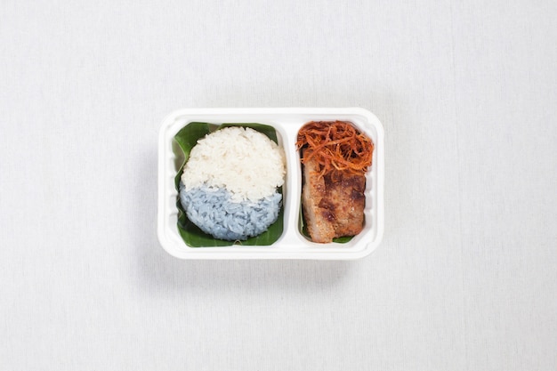 Two color sticky rice with grilled pork and shredded pork put in a white plastic box, put on a white tablecloth, food box, Thai food.