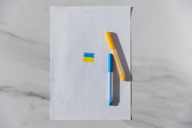 Two colored felttip pens and a symbol in the form of the flag of Ukraine on a white sheet of paper on a white stone background