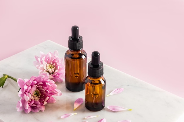 Two cosmetic bottles of amber glass with a pipette with a natural remedy for skin care of the face and body stand on a marble podium with flowers