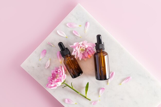 Two cosmetic bottles of dark glass with a natural organic product lie on white marble tiles among the flowers and petals top view pink background