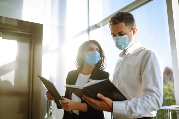 Two coworkers in protective face masks discussing project, sharing ideas.