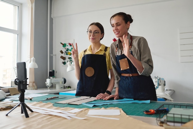 Two creative female tanners in workwear greeting online audience