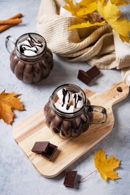 Two cups of aroma hot chocolate with marshmallows and cinnamon on a blue background with autumn maple leaves Concept cozy drink Top view