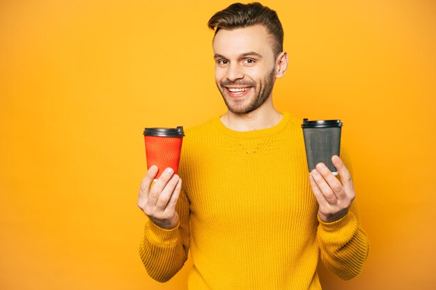 Two cups of coffee or tea in cups of red and black colors in hands of nice-looking man clothed in yellow sweater in front of matching wall