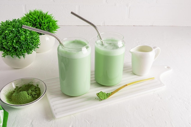 Two cups in the form of a beer can with freshly prepared matcha latte tea on a white ceramic board opposite the brick wall and plants in flower pots