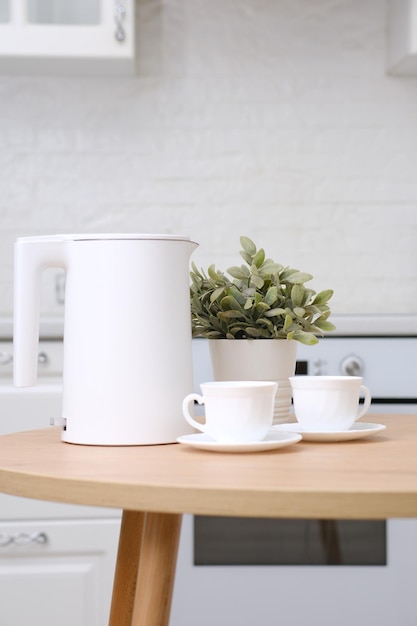 Two cups of tea and white electric kettle green flower in pot on wooden round table in white cosy