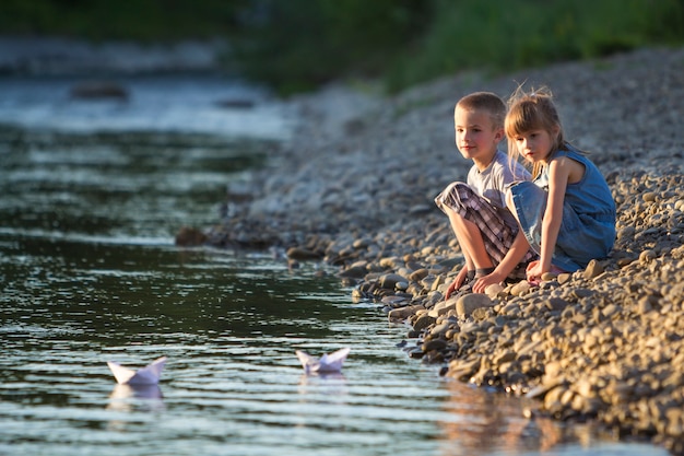 Two cute blond children, boy and girl on river bank sending in water white paper boats on bright summer