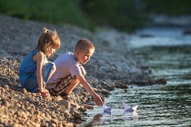 Two cute blond children, boy and girl on river bank sending in water white paper boats 