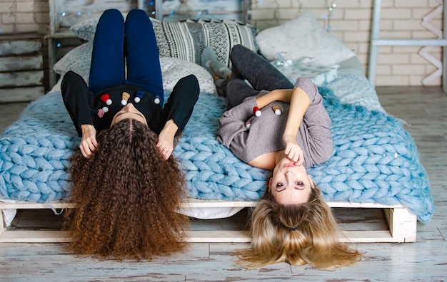 Two cute girlfriends in Christmas jumpers and jeans laying on a warm wrap by head down on the bed in the room