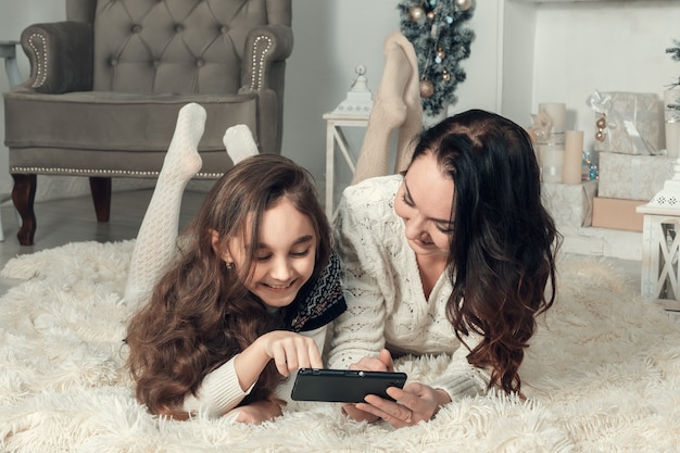 Two cute girls, mother and daughter lying on a floor in Christmas decorated room, use a mobile phone.