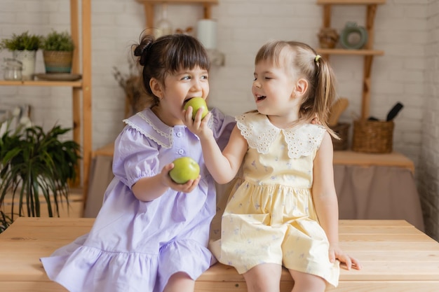 two cute little funny girls in dresses eating green apples in the kitchen. space for text, banner