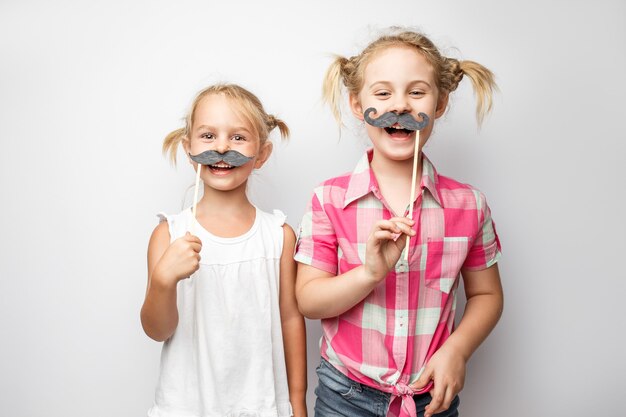 Two cute little girls with paper mustaches while posing against white background