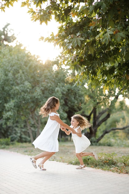 Two cute siblings girls wear similar white dress hoding hands and dance together in park over green nature background outdoor Childhood Friendship