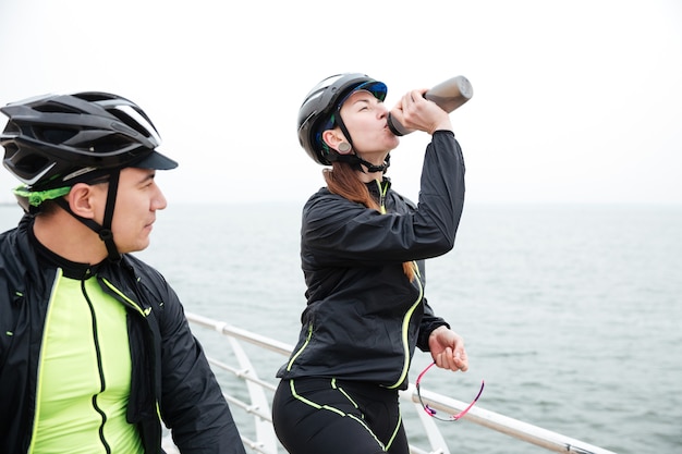 Two cyclists resting near sea. Woman drinking water and man looking at sea