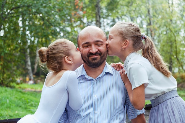 Two daughters with care and gratitude kiss dad on a cheeks and he smiles with happiness on a walk in the park