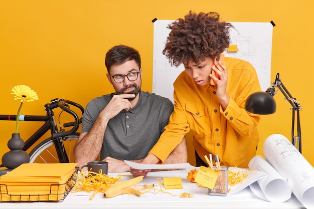 Two diverse architects talk about interior planning and organization of working process pose at coworking space. Puzzled Afro American woman discuss details of cooperation while calling partner