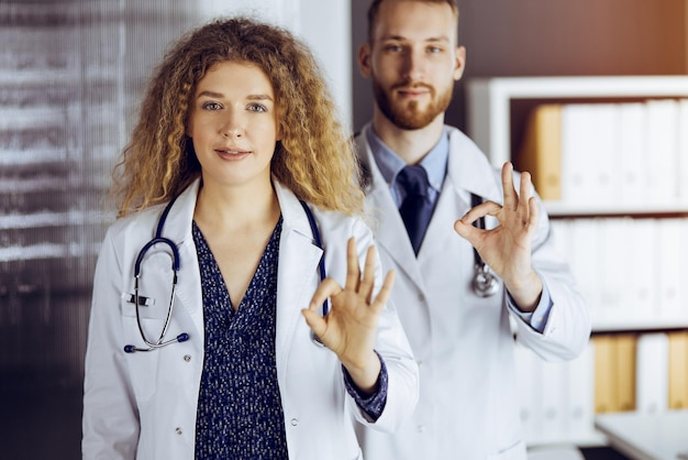 Two doctors standing with thumbs up in hospital office. Medical help, countering viral infection and medicine concept