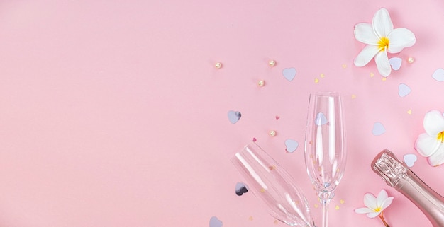 Two empty champagne glasses and bottle of champagne with white frangipani flowers and small heart decoration on pink background, copy space