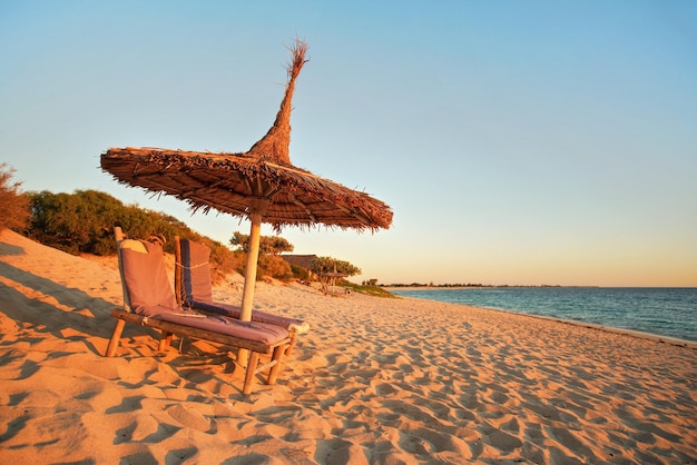 Two empty wooden sunbeds facing the sunset, sea in background, straw sun shade above, image illustrating tropical vacation