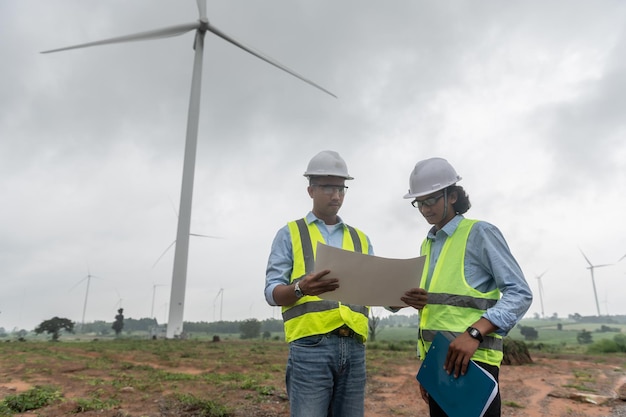 Two engineers working and holding the report at wind turbine farm Power Generator Station on mountainThailand people