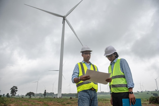 Two engineers working and holding the report at wind turbine farm Power Generator Station on mountainThailand people