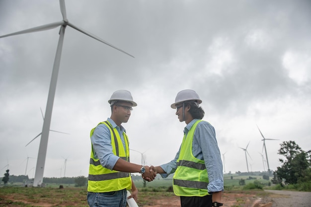 Two engineers working and holding the report at wind turbine farm Power Generator Station on mountainThailand people