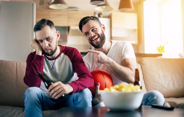 Two excited funny young friends fans of basketball watching TV match and shouting while resting on the couch