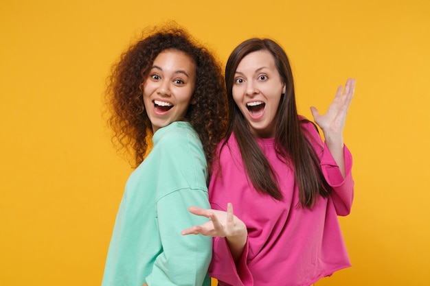 Two excited women friends european and african american girls in pink green clothes posing isolated on yellow orange wall background. People lifestyle concept. Mock up copy space. Spreading hands.