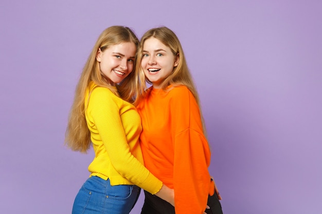 Two excited young blonde twins sisters girls in vivid colorful clothes standing,  isolated on pastel violet blue wall . People family lifestyle concept. 