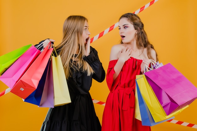 Two excited young women with colorful shopping bags and signal tape isolated over yellow