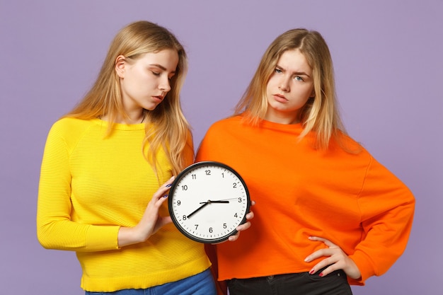 Two exhausted young blonde twins sisters girls in vivid colorful clothes hold round clock isolated on pastel violet blue wall. People family lifestyle concept. 