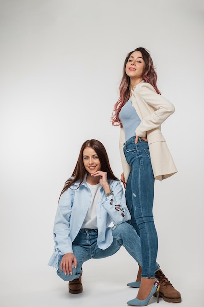 Two fashion pretty woman girlfriends in denim stylish casual outfit sits and poses on a white background in the studio