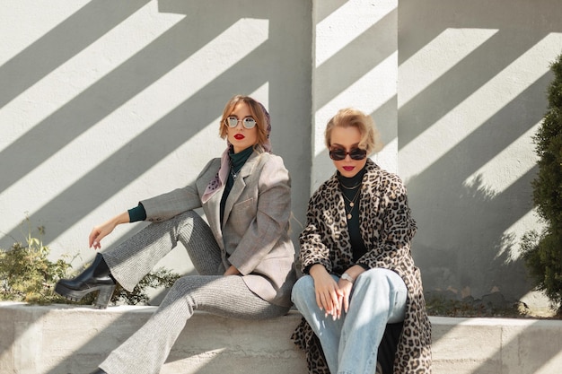 Two fashionable women in stylish retro clothes with leopard print blue jeans shoes coat and sunglasses glasses are sitting near a concrete wall on a sunny day