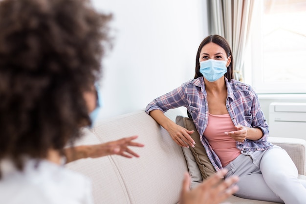 Two female best friends sitting in social distance wearing face mask and talking on the sofa, preventing covid 19 coronavirus infection spread.