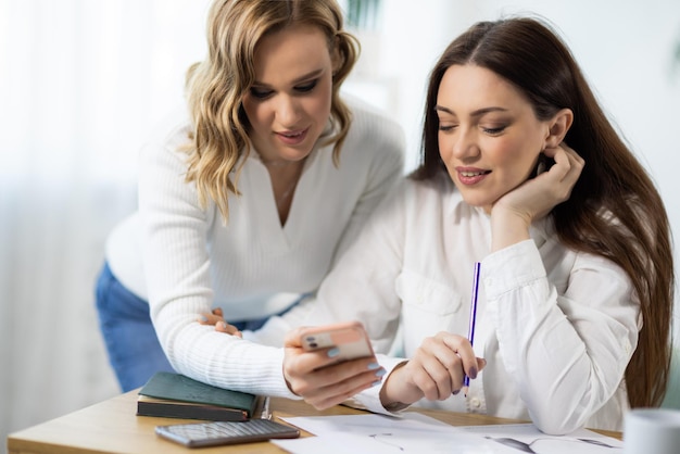 Two female employees are discussing the details of project using information from Internet on phone near table in office