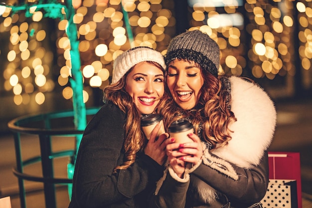 Two female friends enjoy the night out hugging and laughing with cups of coffee in their hands.