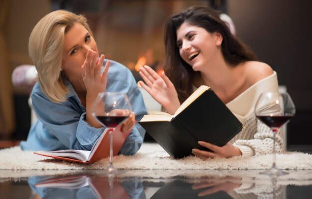 Photo two female friends reading a book and drinking red wine by a fire place. life stile concept.