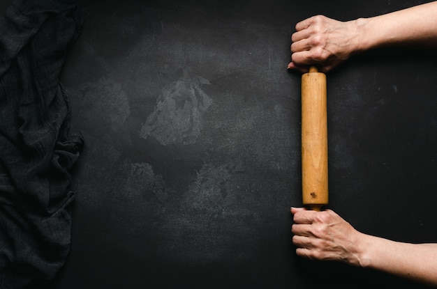 Two female hands hold a wooden rolling pin for rolling dough on a black table top view Place for an inscription