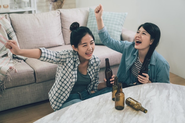 two female happy friends football fans watching soccer on tv and celebrating victory at home. Friendship sports and entertainment concept. asian women sitting on wooden floor drinking bottles of beer