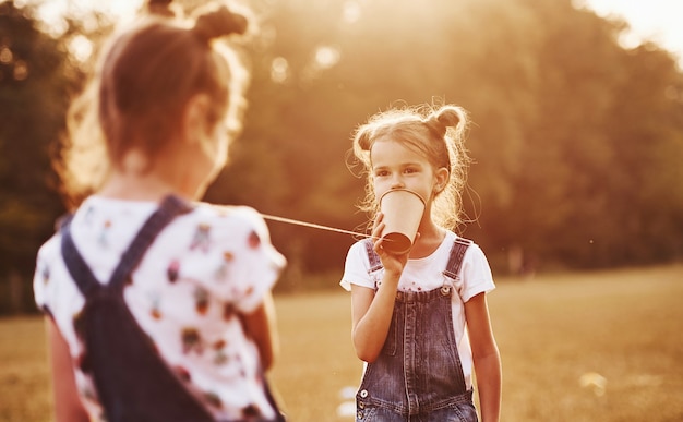 Two female kids stands in the field and talking by using string can phone.