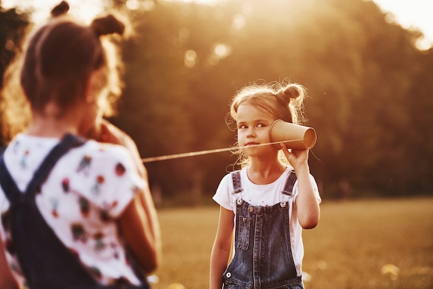 Two female kids stands in the field and talking by using string can phone.